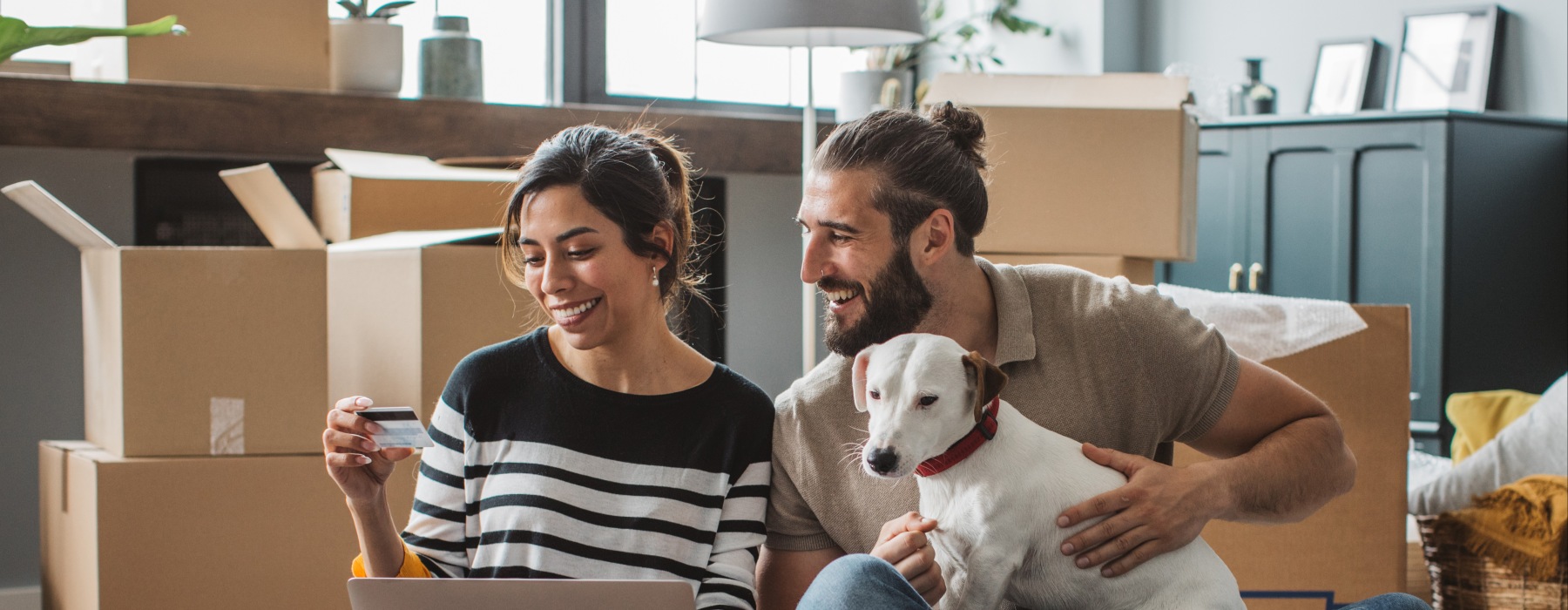 A smiling couple sitting on the floor of their new pet-friendly apartment surrounded by moving boxes, using a laptop and credit card with their dog.